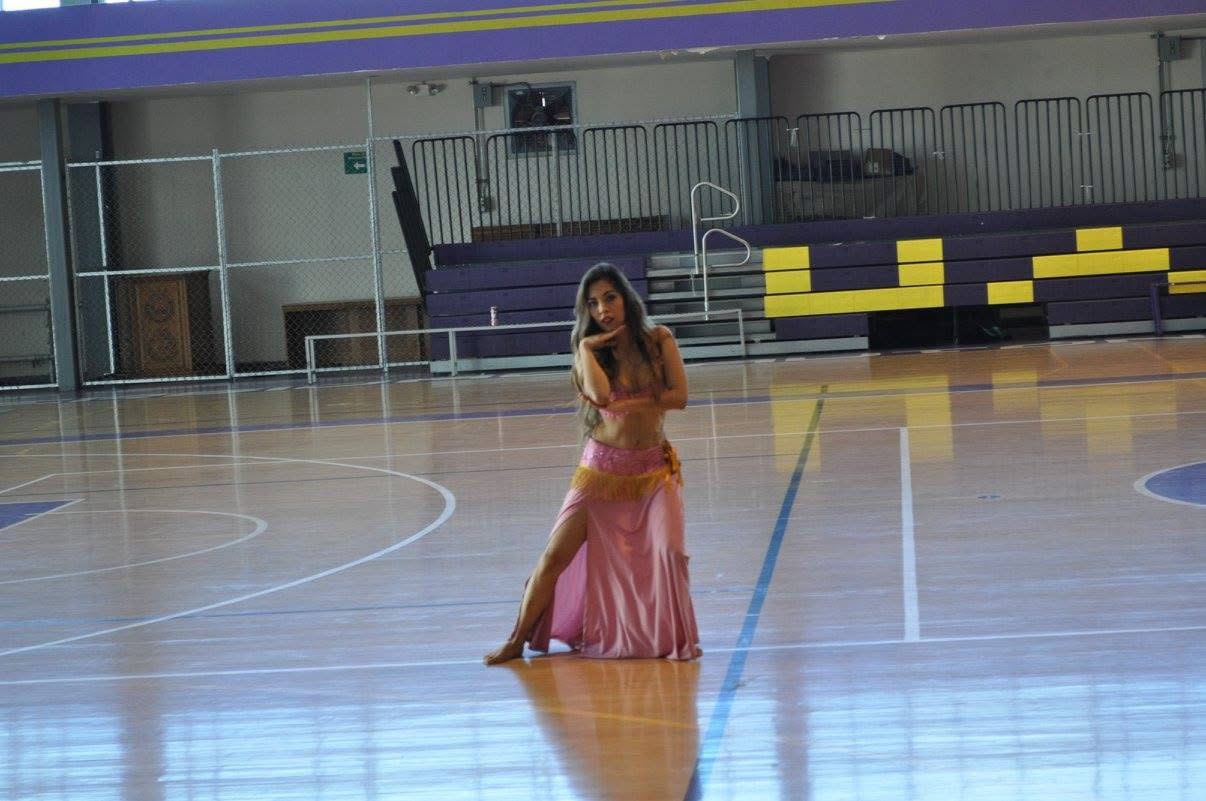 Una bailarina con traje rosa de danza árabe posa arrodillada sobre la duela brillante de un gimnasio universitario con gradas moradas y amarillas.