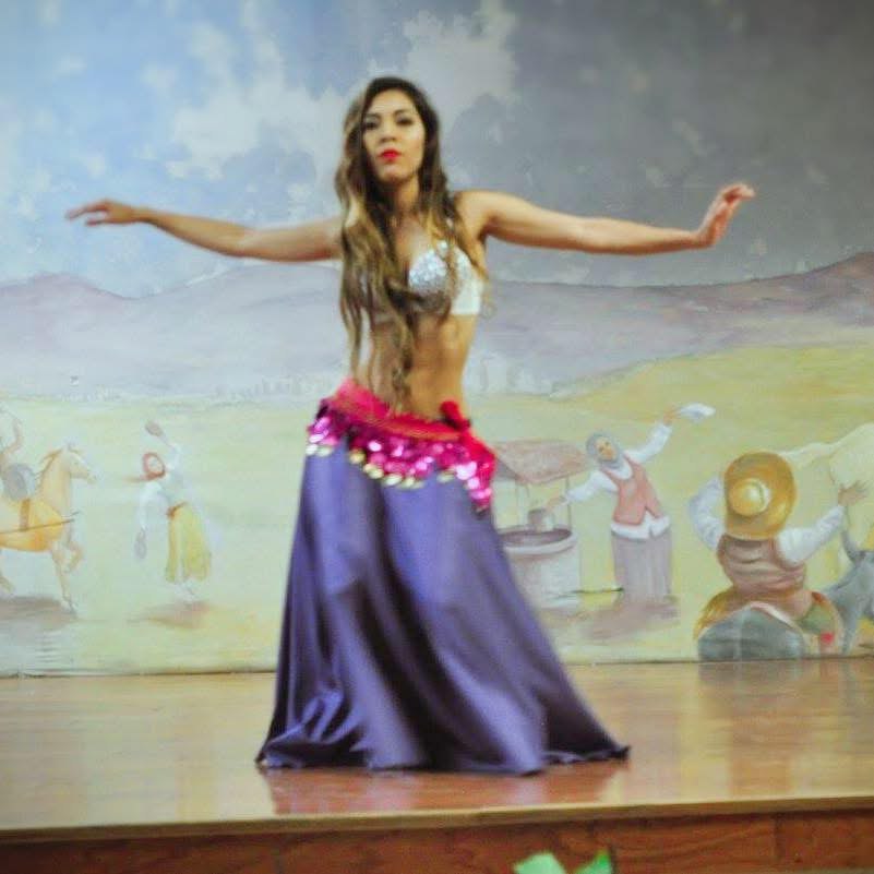 Una bailarina de danza árabe con falda morada y cinturón fucsia abre los brazos sobre un escenario con un mural campestre de fondo.