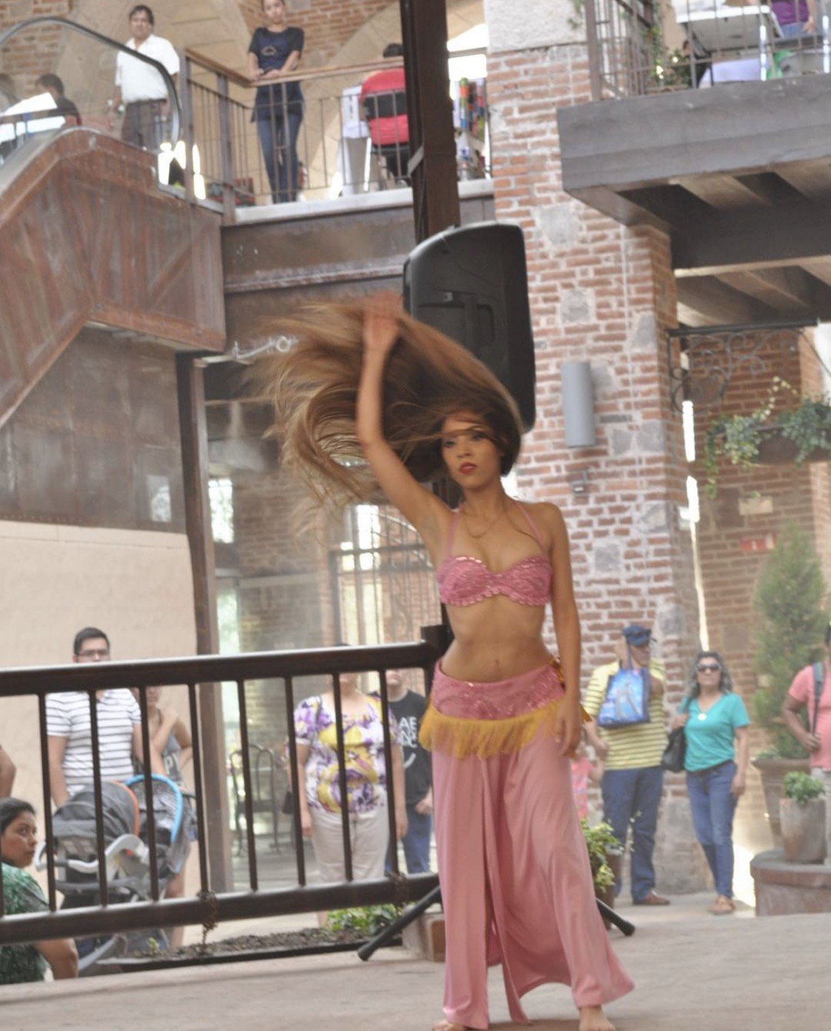 La Dra. Pamela Corona baila danza árabe con traje rosa, agitando su larga melena, ante el público de una plaza de ladrillo en Chihuahua.
