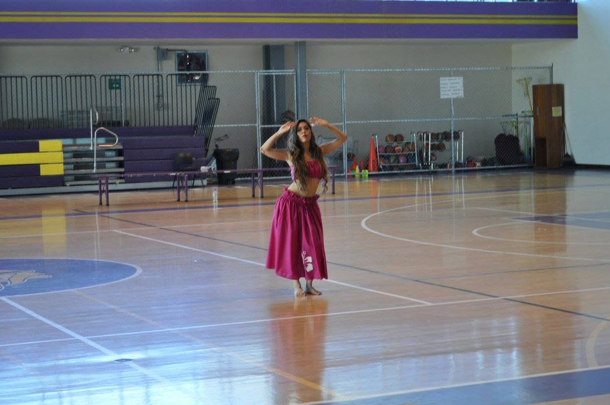 Una bailarina con falda y top fucsia de danza hawaiana, con las manos sobre la cabeza, en medio de un gimnasio universitario vacío.