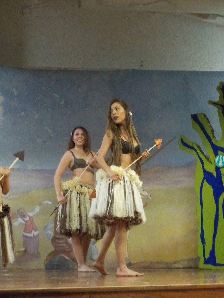 Bailarinas con faldas de fibra natural y lanzas en las manos se mueven sobre un escenario frente a un mural y un árbol pintado en verde y azul.