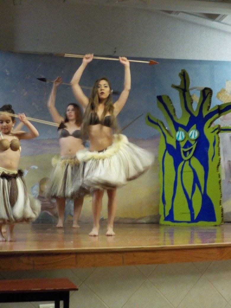 Tres bailarinas con faldas de fibra natural levantan lanzas sobre la cabeza en un escenario, junto a un árbol pintado en verde y azul.