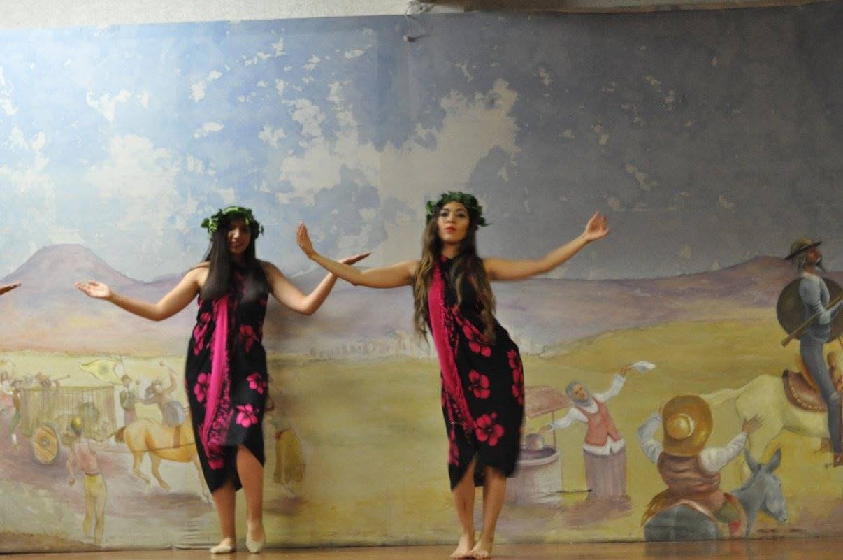 Dos bailarinas con pareos negros de flores fucsia y coronas de hojas bailan con los brazos abiertos frente a un mural de paisaje.