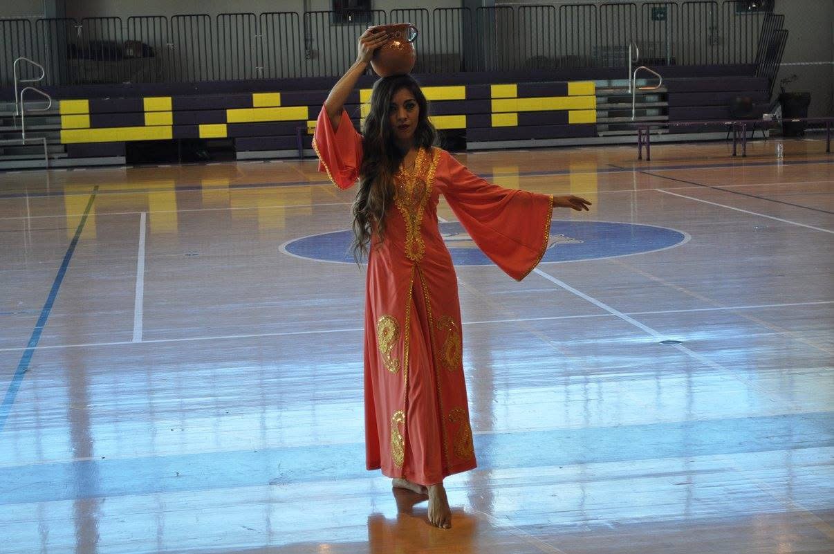 Una bailarina con túnica coral bordada en dorado equilibra una vasija de barro sobre la cabeza en la duela de un gimnasio universitario.