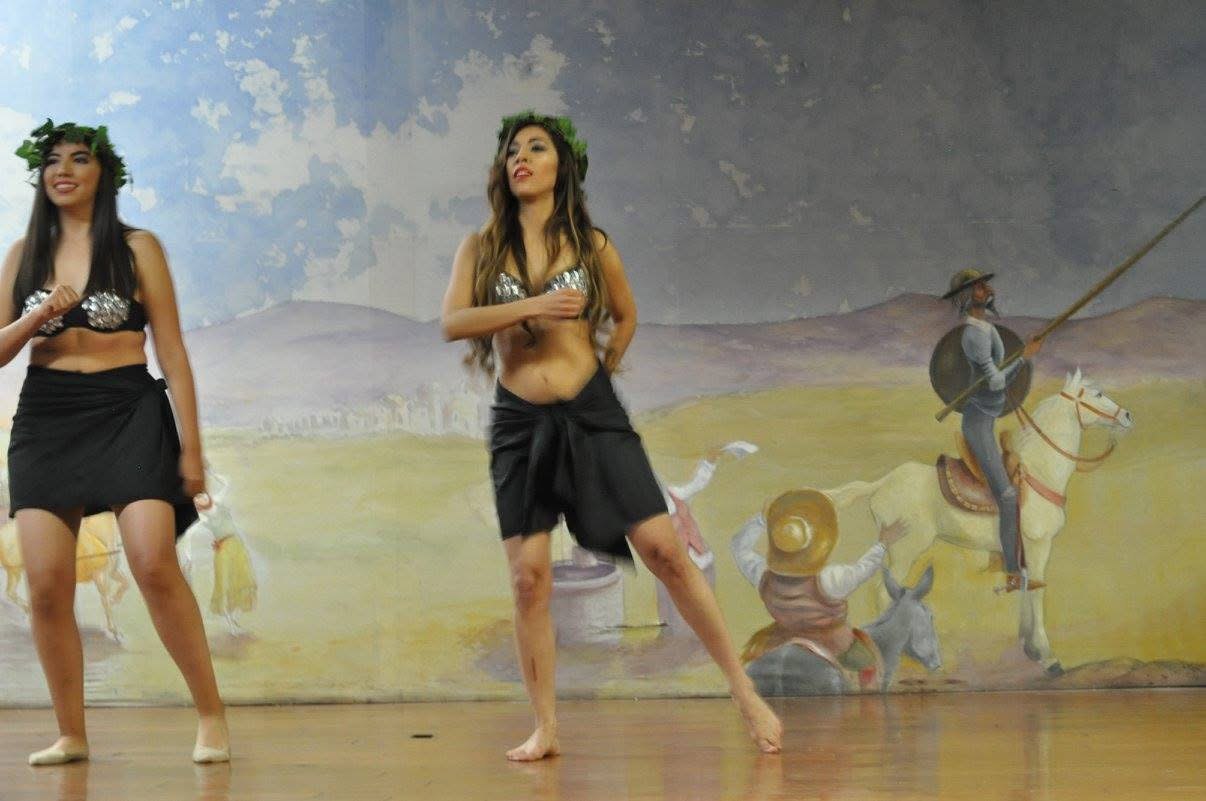 Dos bailarinas de danza tahitiana con top plateado, pareo negro y corona de hojas bailan sobre un escenario frente a un mural del Quijote.