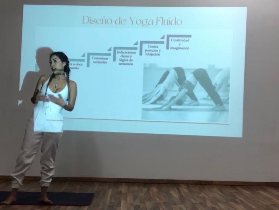 Una mujer vestida de blanco explica frente a una proyección titulada «Diseño de Yoga Fluido» con los pasos para diseñar una secuencia.