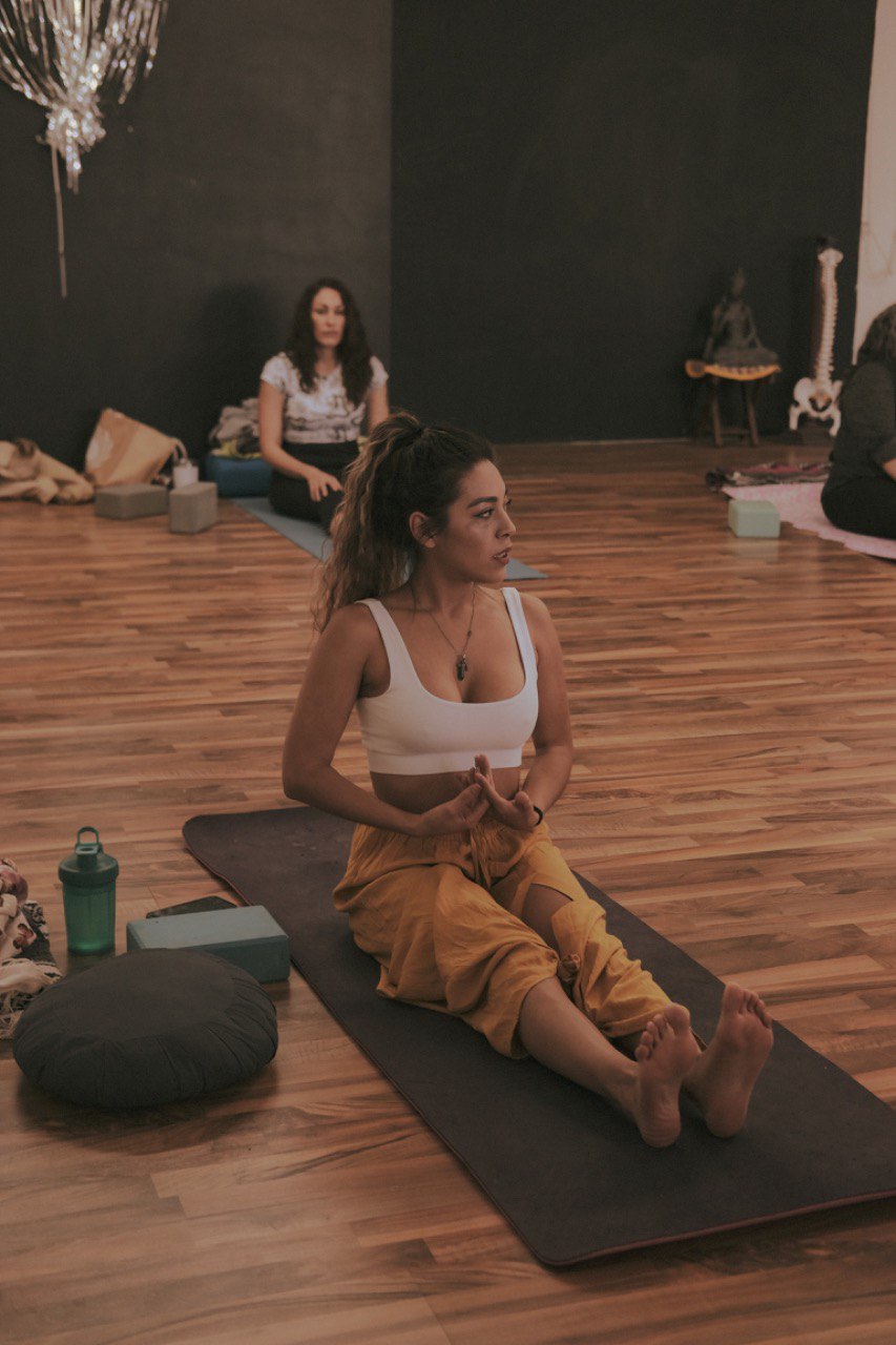 Una mujer con top blanco y pantalón mostaza, sentada con las piernas extendidas sobre un tapete, habla al grupo en un salón con piso de madera.