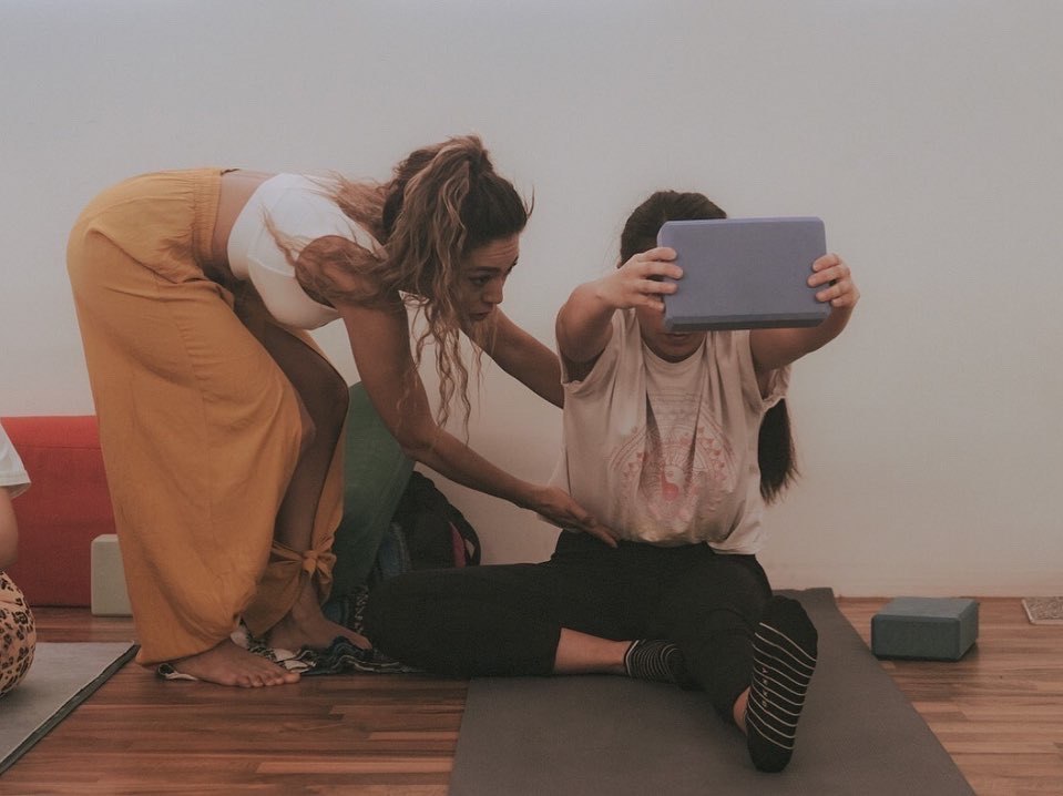 La instructora, con pantalón mostaza, se inclina para ajustar la postura de una alumna sentada que sostiene un bloque de yoga con los brazos extendidos.