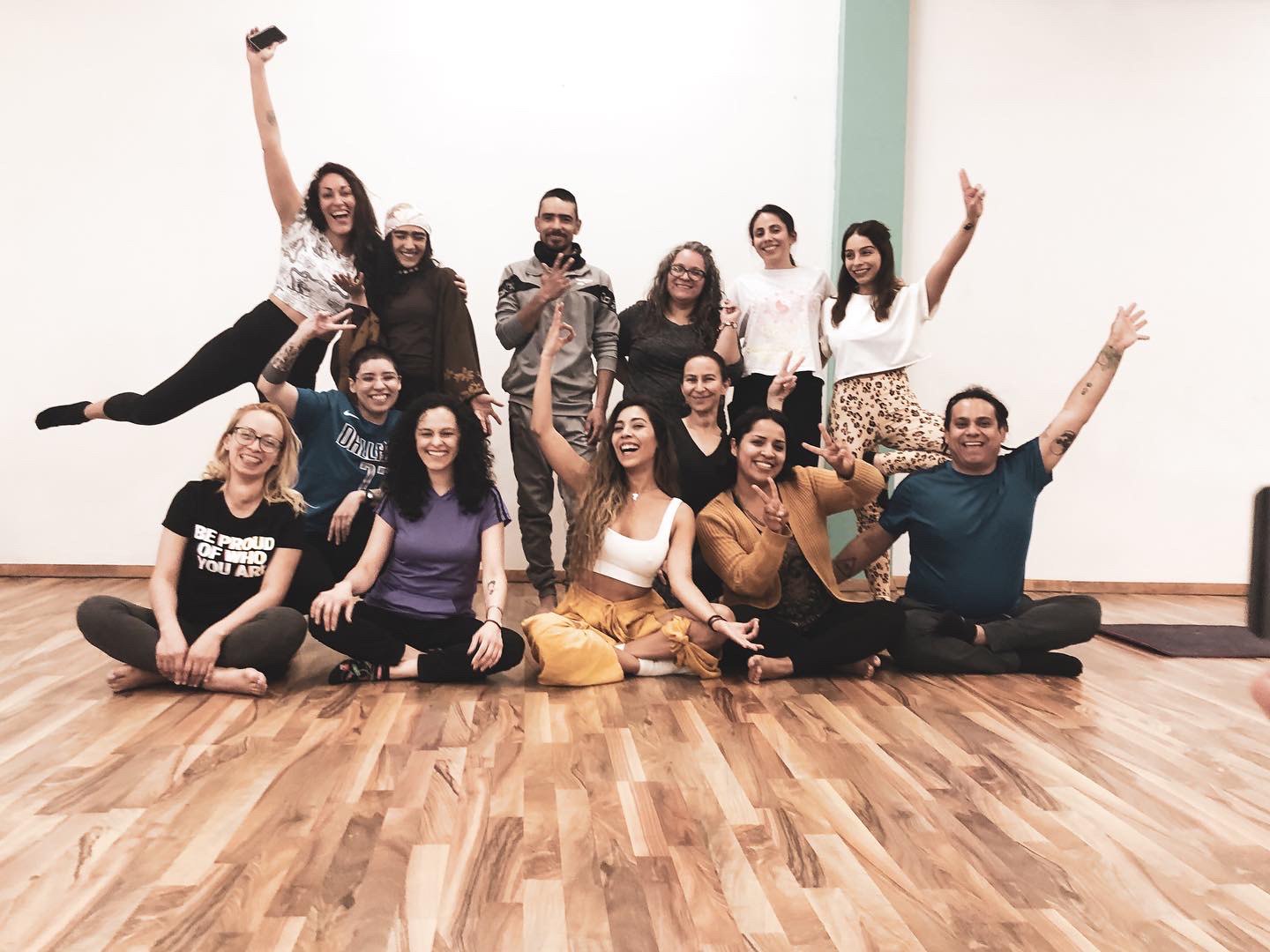 Foto de grupo alegre de unas quince personas en un estudio de yoga con piso de madera, algunas sentadas y otras de pie con los brazos en alto.