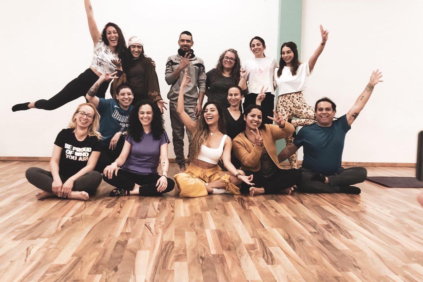 Foto de grupo alegre de unas quince personas en un estudio de yoga con piso de madera, algunas sentadas y otras de pie con los brazos en alto.