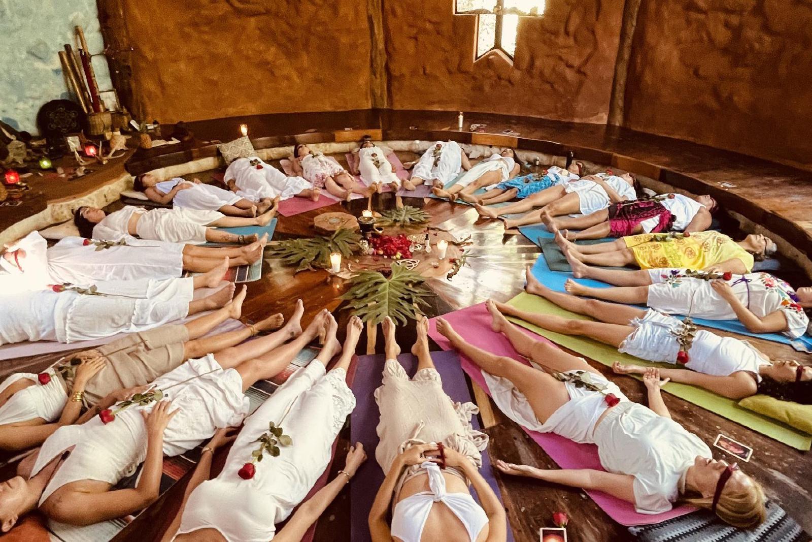 Mujeres vestidas de blanco yacen en círculo sobre tapetes de colores alrededor de un altar con velas y hojas, cada una con una rosa roja, en un espacio de barro.
