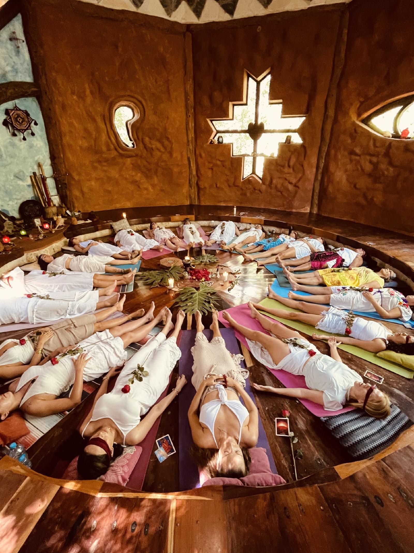 Mujeres vestidas de blanco yacen en círculo sobre tapetes de colores alrededor de un altar con velas y hojas, cada una con una rosa roja, en un espacio de barro.