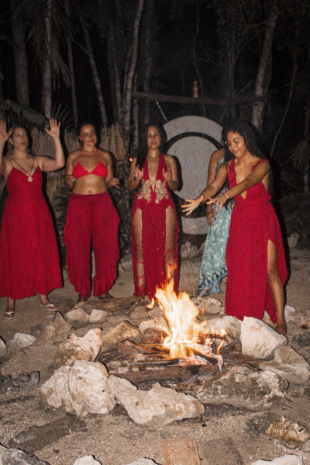 De noche, en la selva, mujeres vestidas de rojo bailan alrededor de una fogata rodeada de piedras, con un gong detrás.