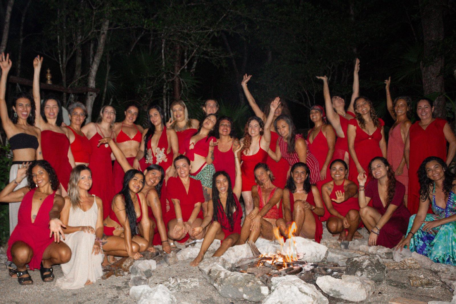 Retrato de grupo nocturno de unas treinta mujeres vestidas de rojo, sonrientes y con los brazos en alto alrededor de una fogata en la selva.