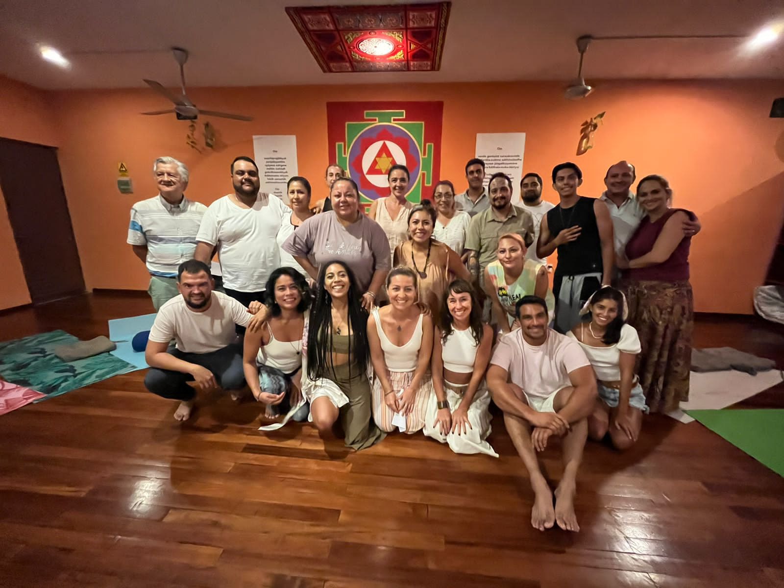 Retrato de grupo de unas veinticinco personas, mujeres y hombres, sonrientes en una sala naranja con un yantra rojo y verde en la pared.