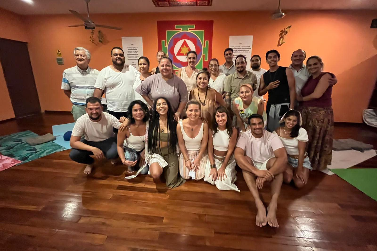 Retrato de grupo de unas veinticinco personas, mujeres y hombres, sonrientes en una sala naranja con un yantra rojo y verde en la pared.