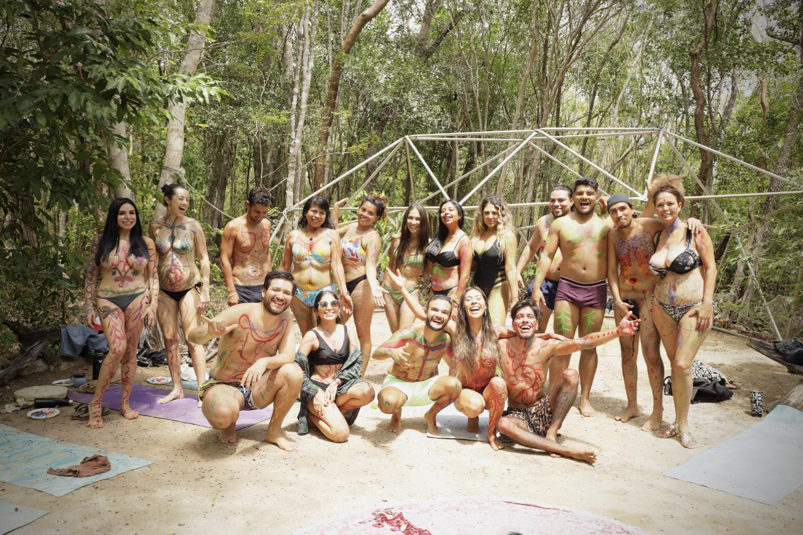 Retrato de grupo de mujeres y hombres con el cuerpo pintado de colores, en traje de baño, bajo una estructura de domo en la selva.