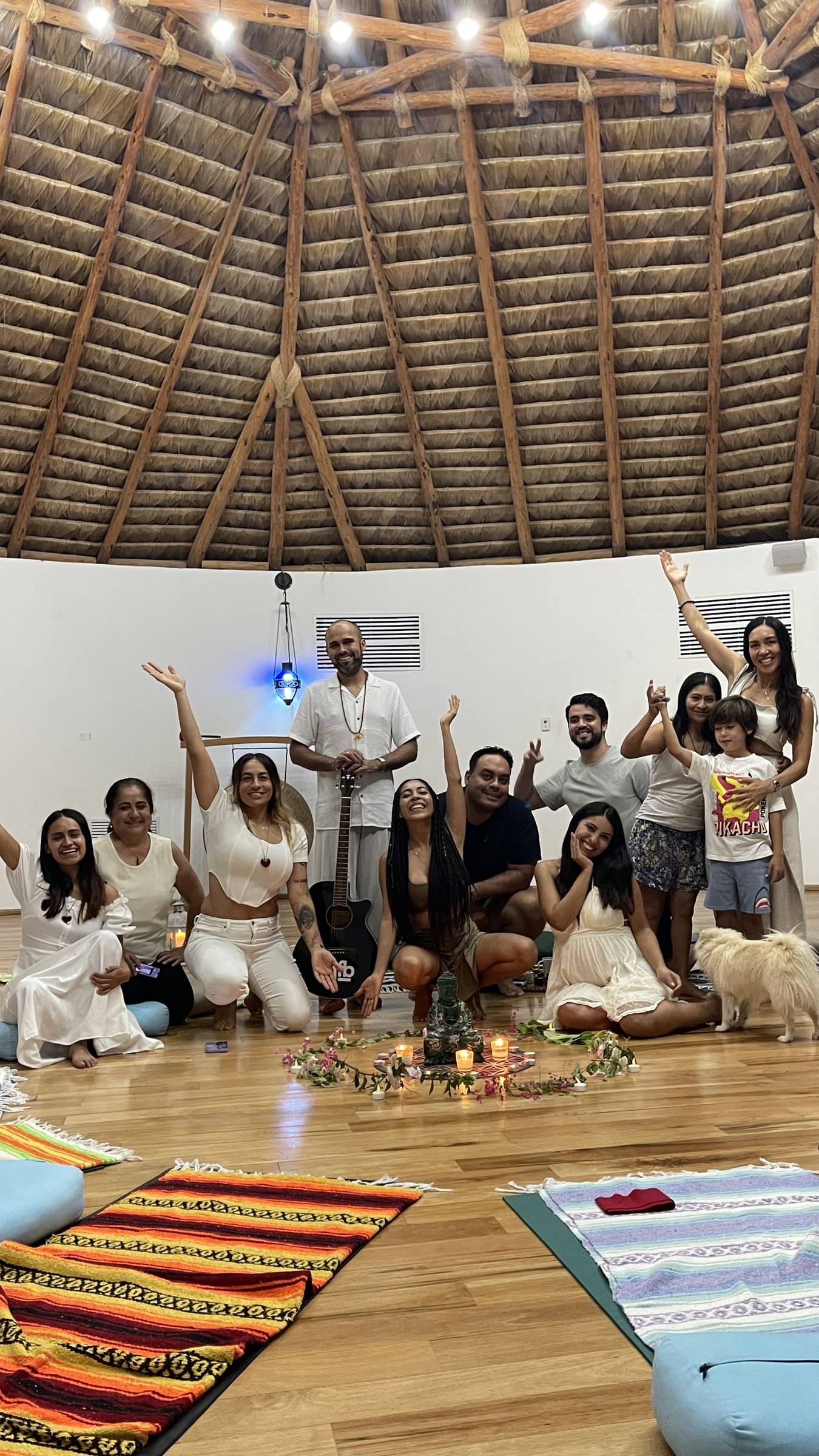 Un grupo sonriente, casi todo de blanco, reunido con un músico con guitarra alrededor de un altar de velas y flores bajo un techo de palma.