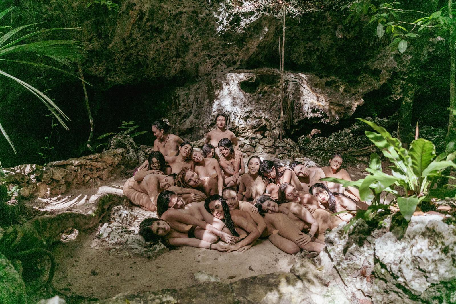 Un grupo numeroso de mujeres desnudas, recostadas unas sobre otras en la arena bajo una bóveda de roca, rodeadas de selva y luz moteada.