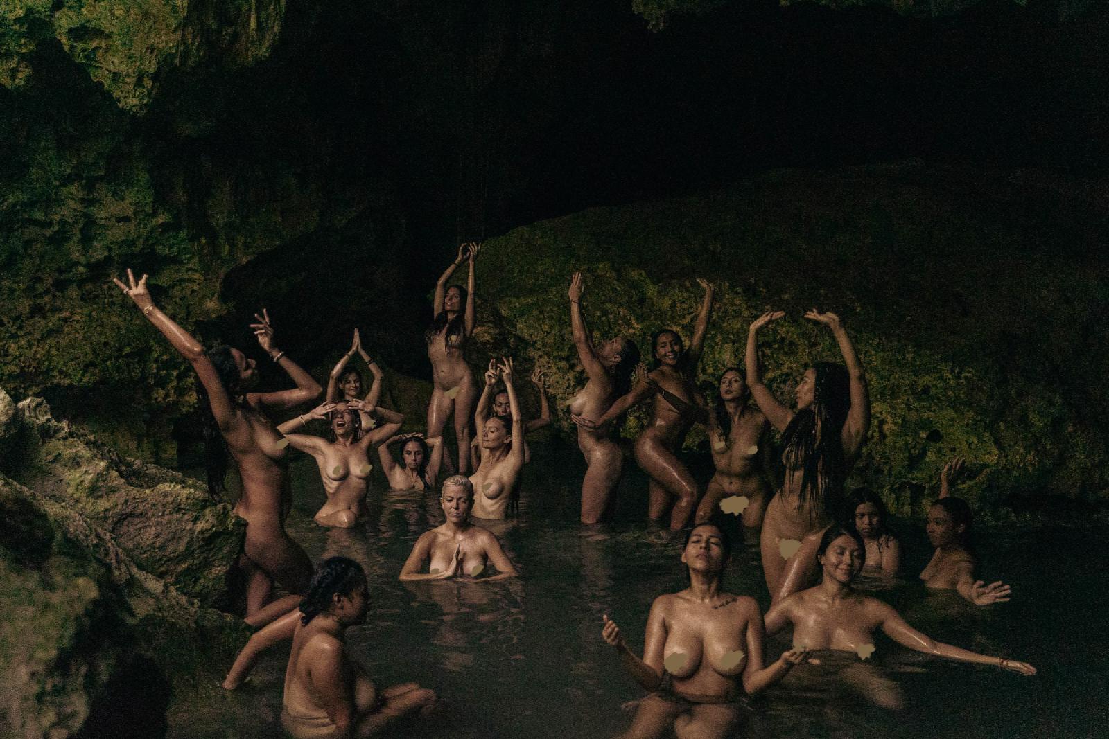 Un grupo de mujeres desnudas en el agua de un cenote en penumbra, algunas con los brazos alzados y otras con las manos juntas.