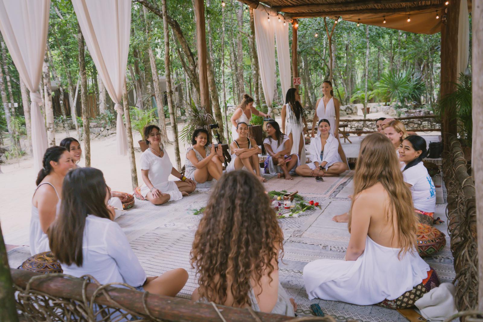 Mujeres vestidas de blanco sentadas en círculo sobre alfombras bajo una palapa con cortinas, alrededor de un altar de flores y hierbas, en la selva.