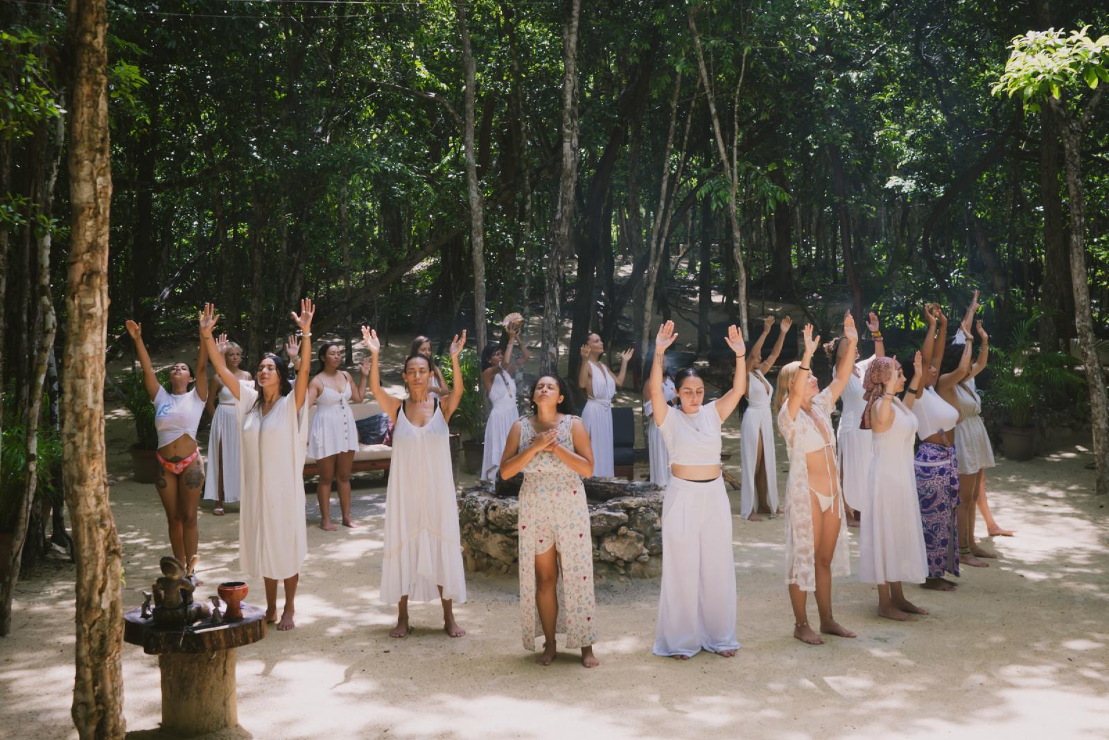Mujeres vestidas casi todas de blanco, descalzas sobre arena en la selva, con los ojos cerrados y los brazos alzados alrededor de un círculo de piedra.