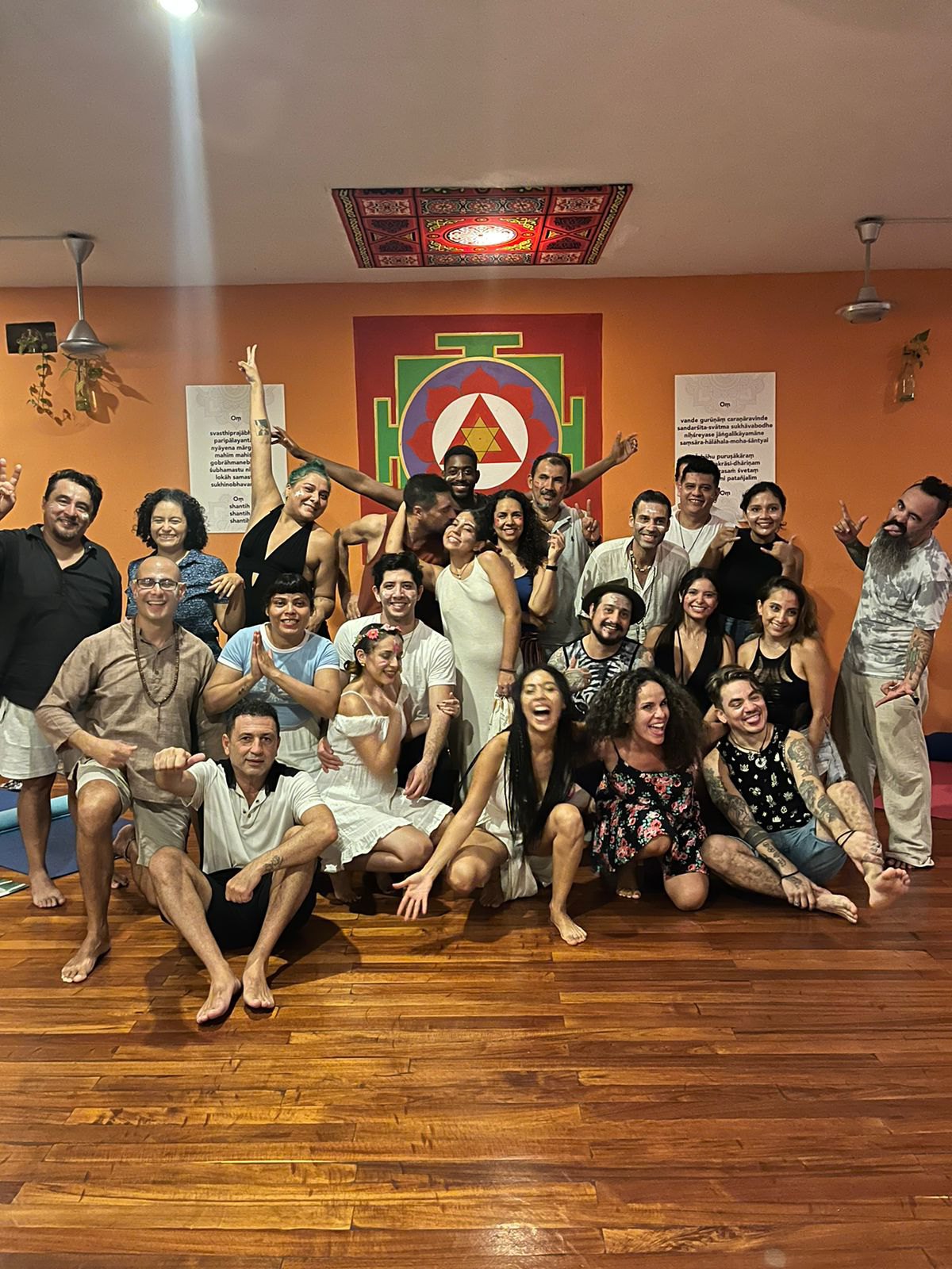 Foto de grupo sonriente de hombres y mujeres en un salón de yoga con paredes naranjas, un gran yantra pintado y suelo de madera.