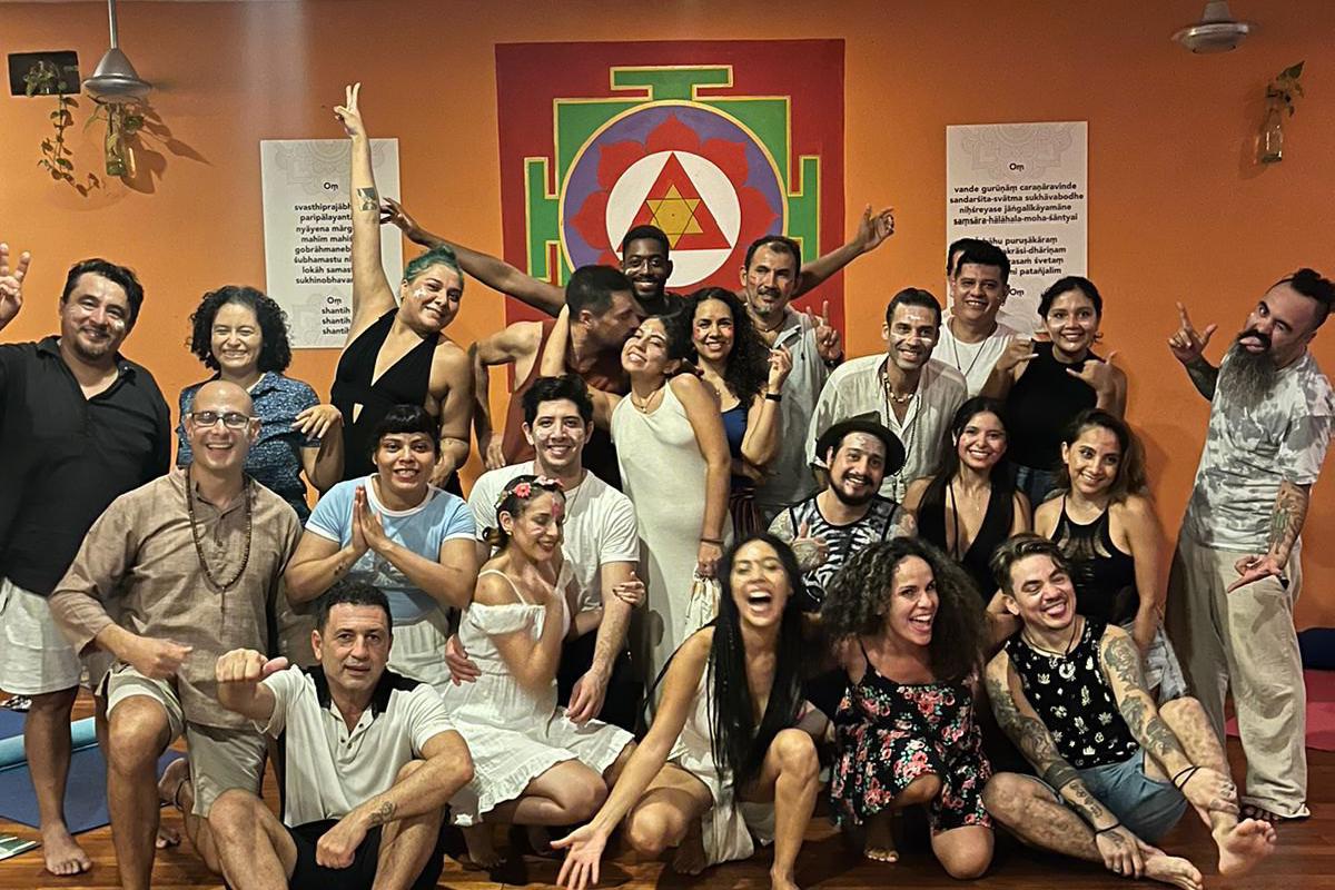 Foto de grupo sonriente de hombres y mujeres en un salón de yoga con paredes naranjas, un gran yantra pintado y suelo de madera.