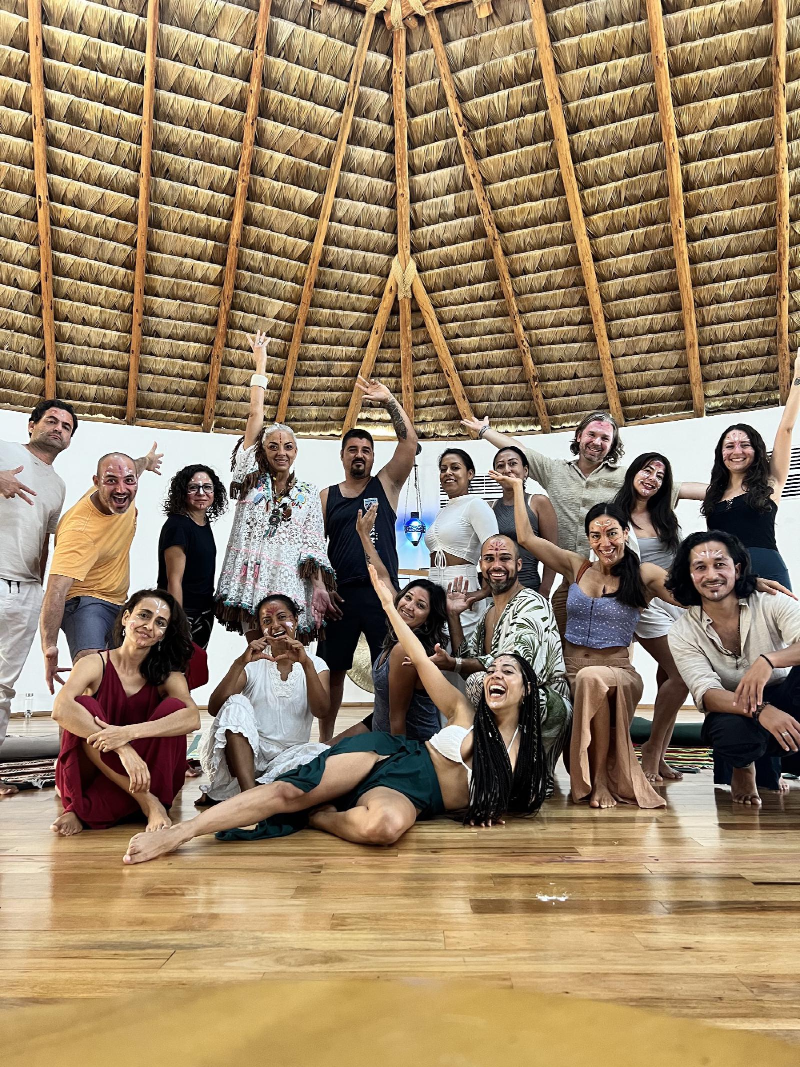 Grupo de hombres y mujeres con pintura blanca en el rostro, posando alegres bajo el techo de palma de una gran palapa circular con piso de madera.