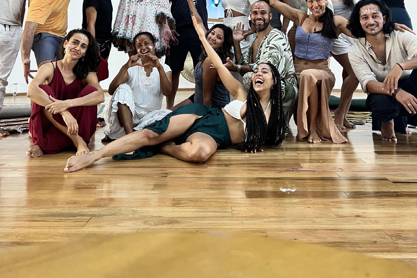 Grupo de hombres y mujeres con pintura blanca en el rostro, posando alegres bajo el techo de palma de una gran palapa circular con piso de madera.