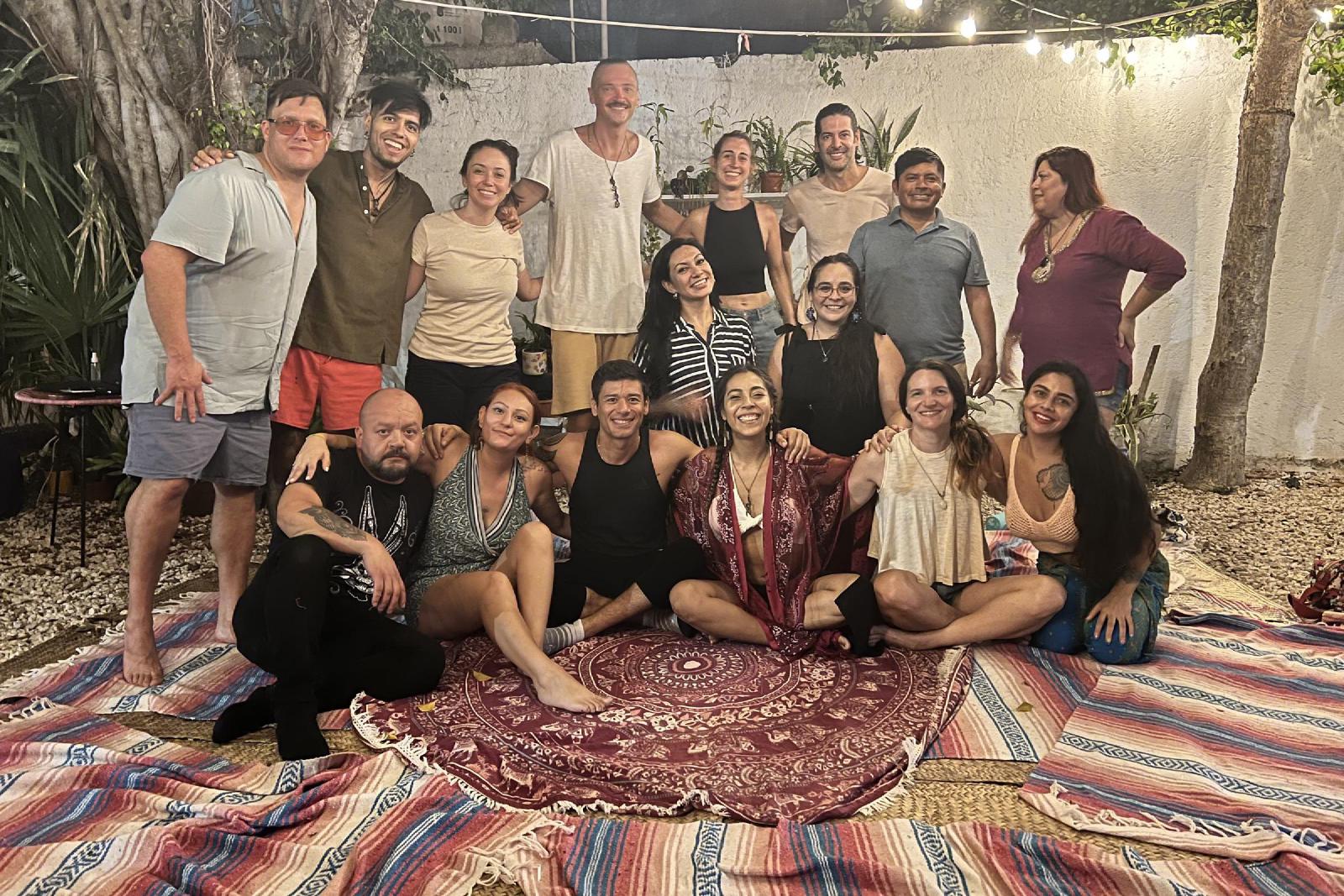 Foto de grupo de noche en un patio con luces colgantes, hombres y mujeres sonrientes sentados y de pie sobre sarapes y un tapete de mandala.