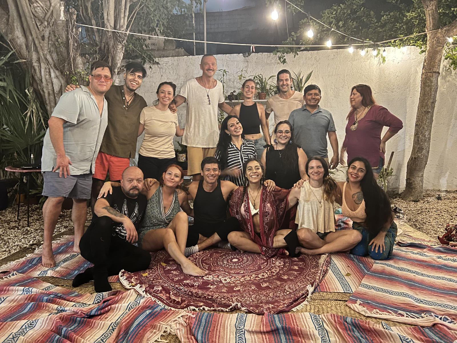 Foto de grupo de noche en un patio con luces colgantes, hombres y mujeres sonrientes sentados y de pie sobre sarapes y un tapete de mandala.