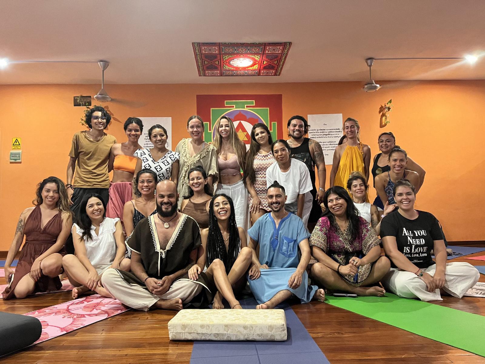 Foto de grupo sonriente de unas veinticinco personas sentadas y de pie en un salón de yoga naranja con yantra pintado y tapetes de colores.
