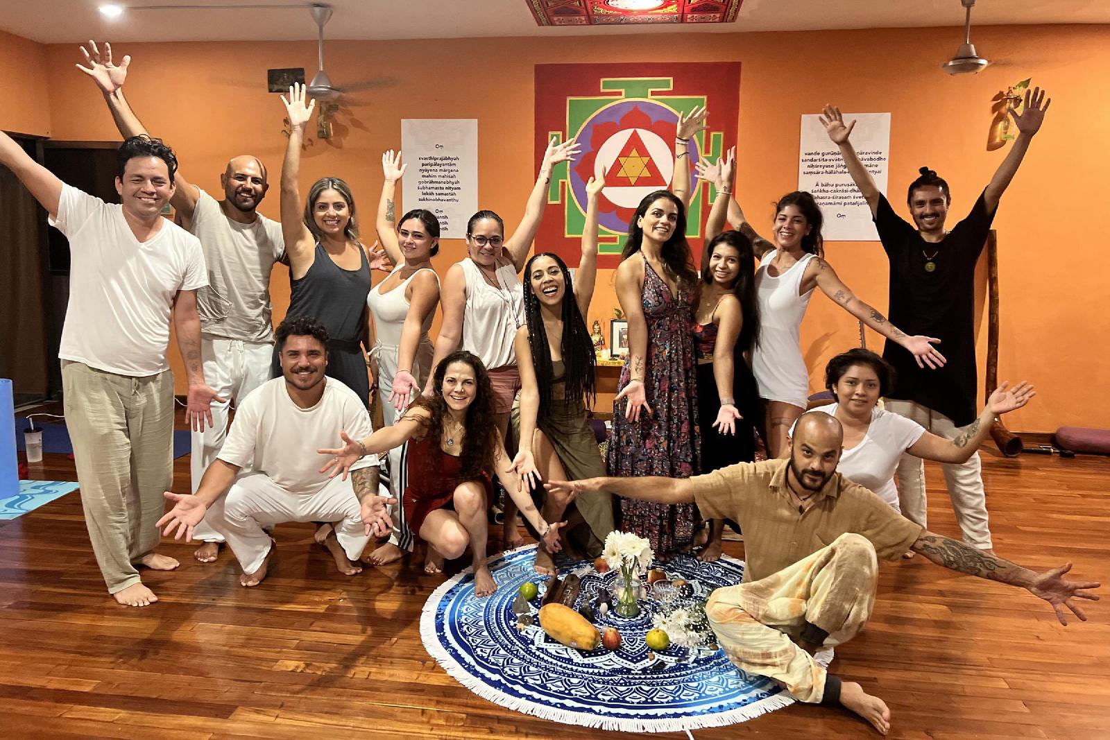 Grupo de hombres y mujeres con los brazos abiertos en un salón de paredes naranjas con yantra pintado, alrededor de un altar de flores y frutas.