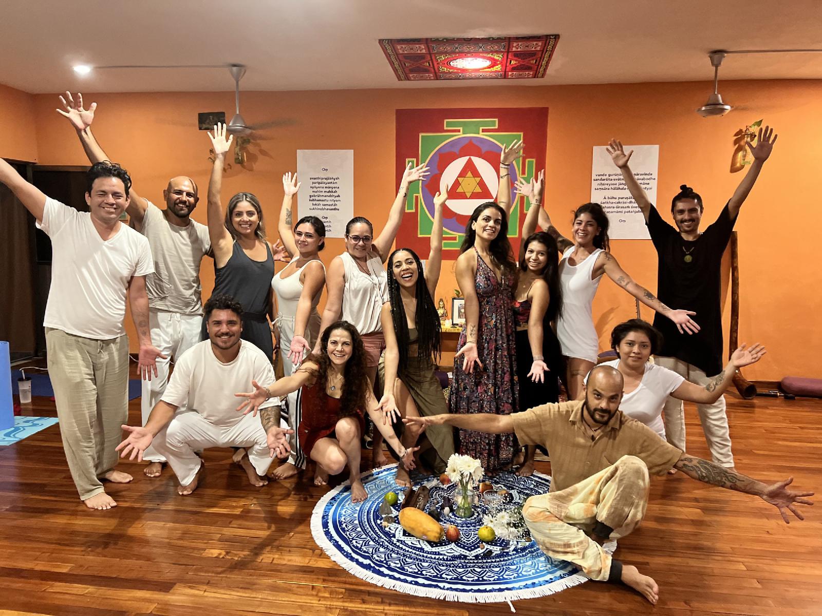 Grupo de hombres y mujeres con los brazos abiertos en un salón de paredes naranjas con yantra pintado, alrededor de un altar de flores y frutas.