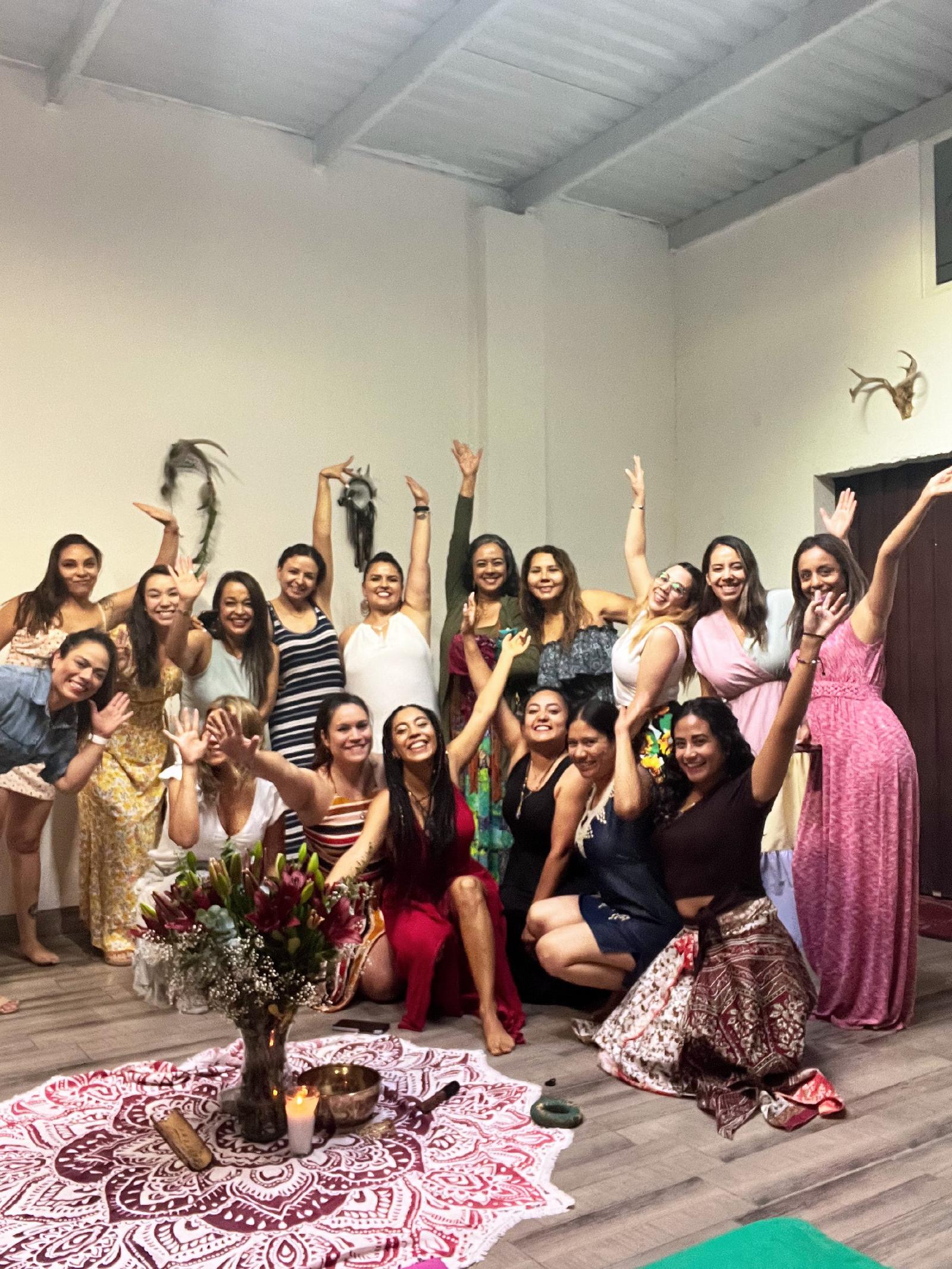 Grupo de mujeres con vestidos de colores, sonrientes y con los brazos en alto, en una sala blanca junto a un altar con flores, vela y cuenco.