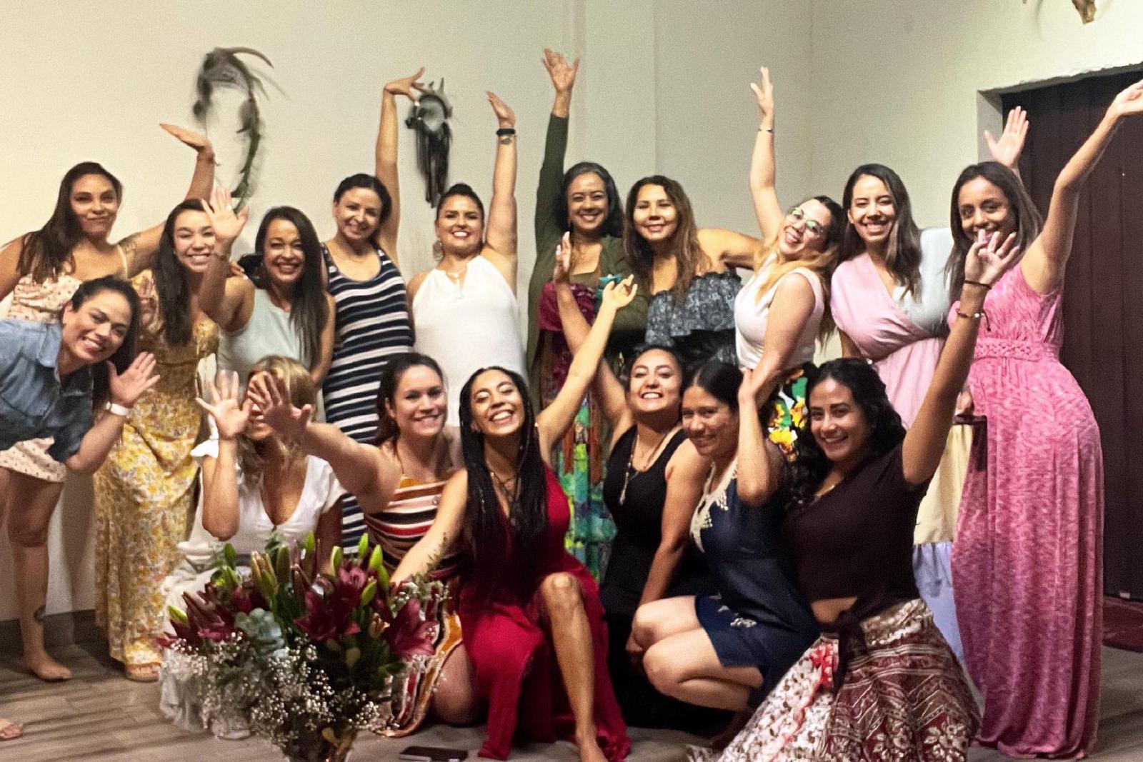 Grupo de mujeres con vestidos de colores, sonrientes y con los brazos en alto, en una sala blanca junto a un altar con flores, vela y cuenco.