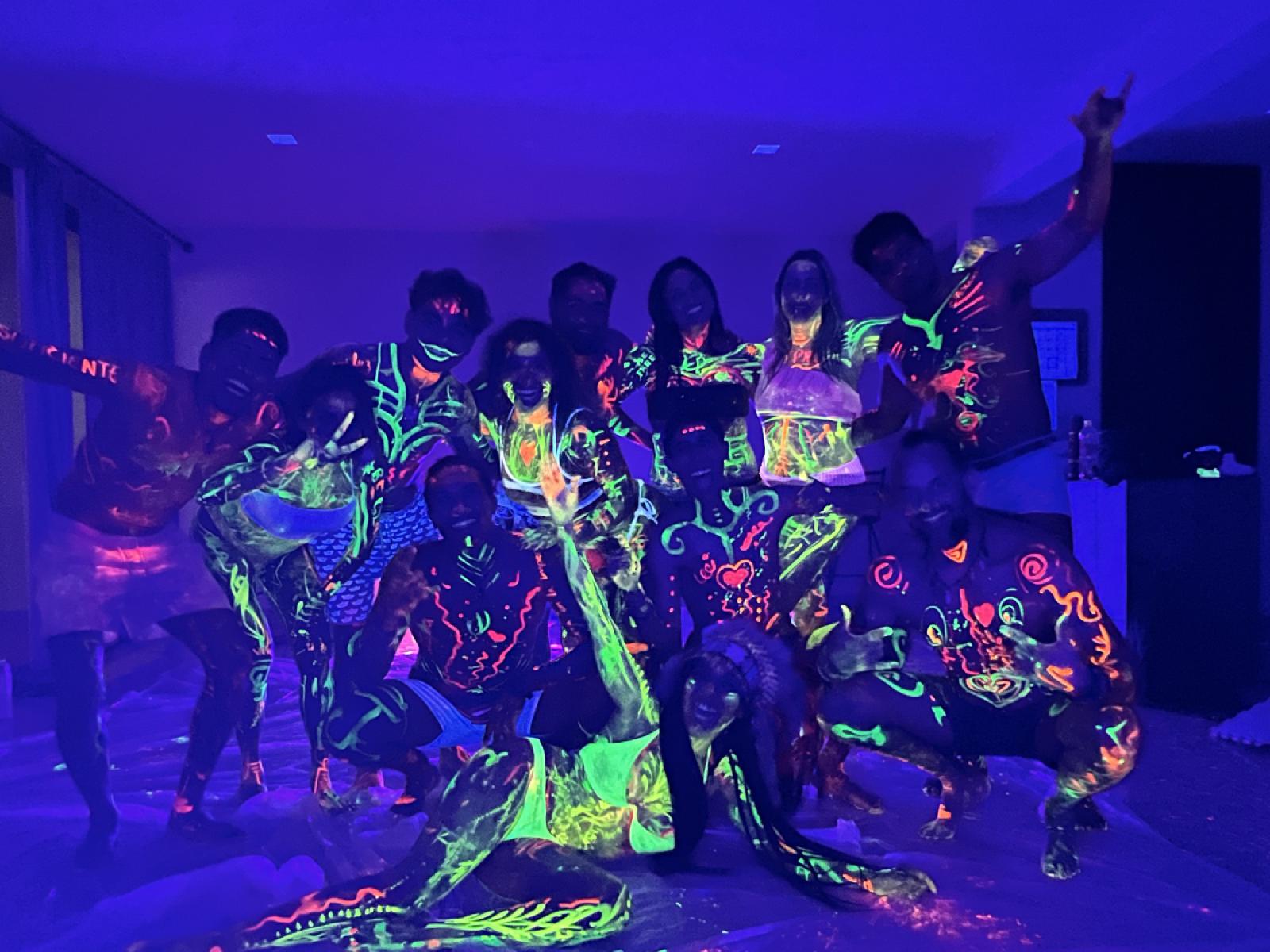 Un grupo de mujeres y hombres con el cuerpo pintado con pintura fluorescente brilla bajo luz ultravioleta en una sala oscura.