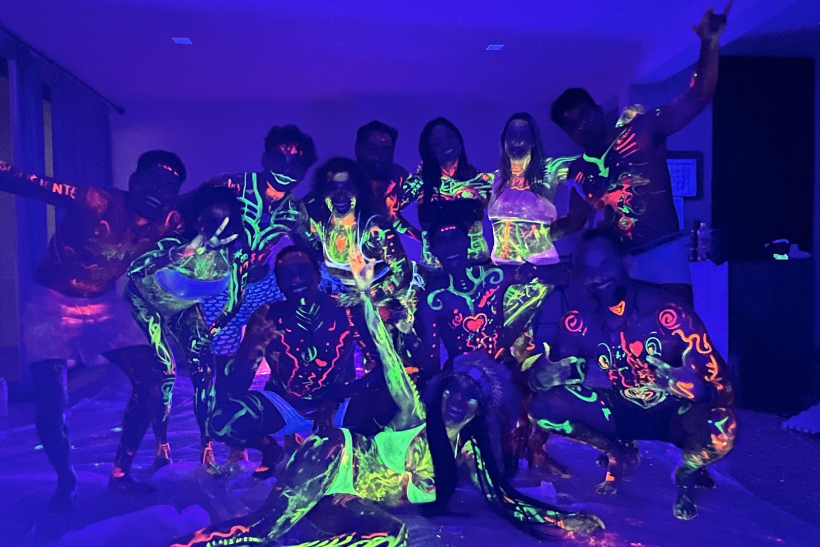 Un grupo de mujeres y hombres con el cuerpo pintado con pintura fluorescente brilla bajo luz ultravioleta en una sala oscura.