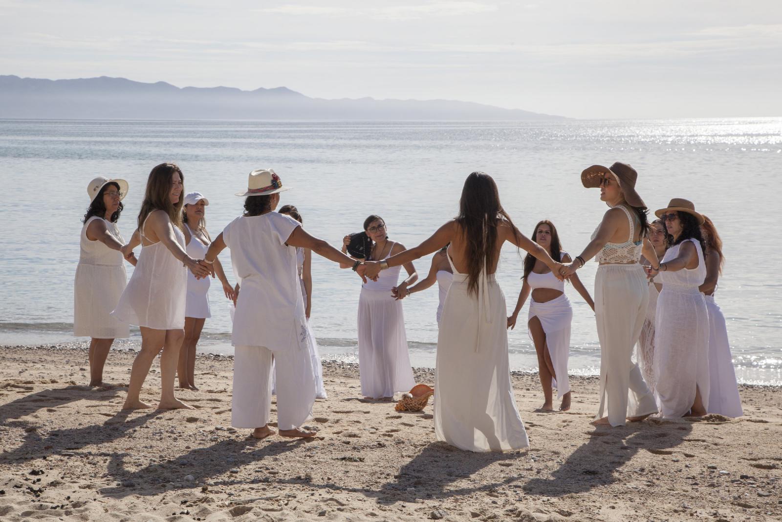 Mujeres vestidas de blanco se toman de las manos formando un círculo en la playa, con el mar en calma y montañas al fondo.