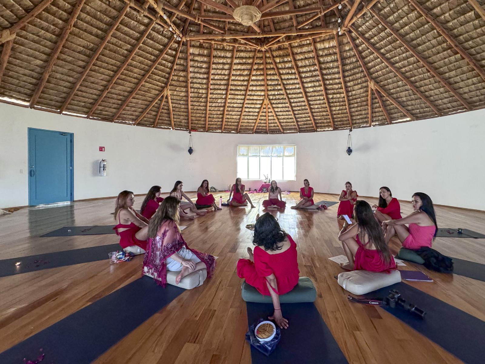 Un grupo de mujeres vestidas de rojo y rosa sentadas en círculo sobre cojines y tapetes, bajo un gran techo de palapa en una sala luminosa.
