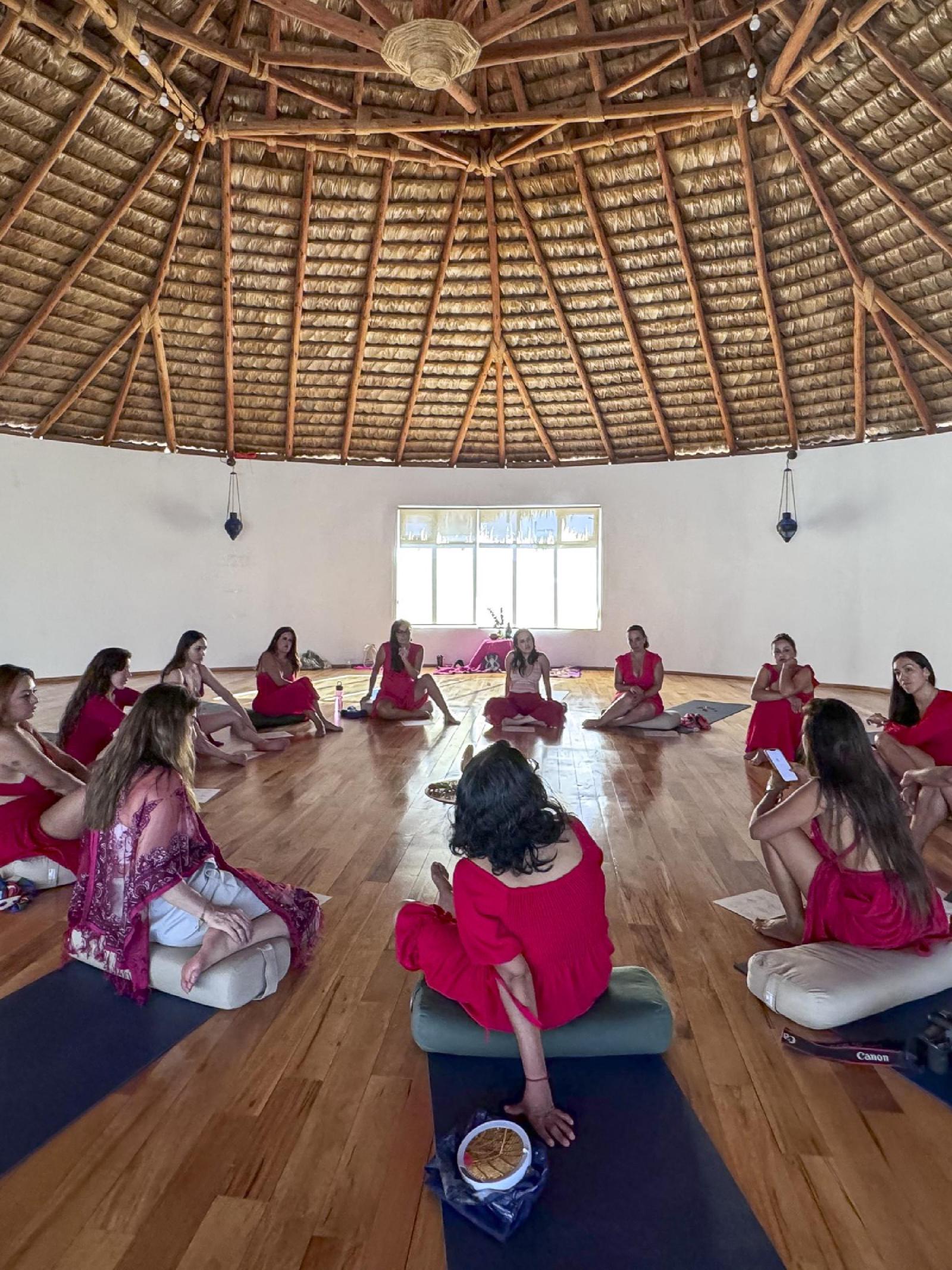 Un grupo de mujeres vestidas de rojo y rosa sentadas en círculo sobre cojines y tapetes, bajo un gran techo de palapa en una sala luminosa.