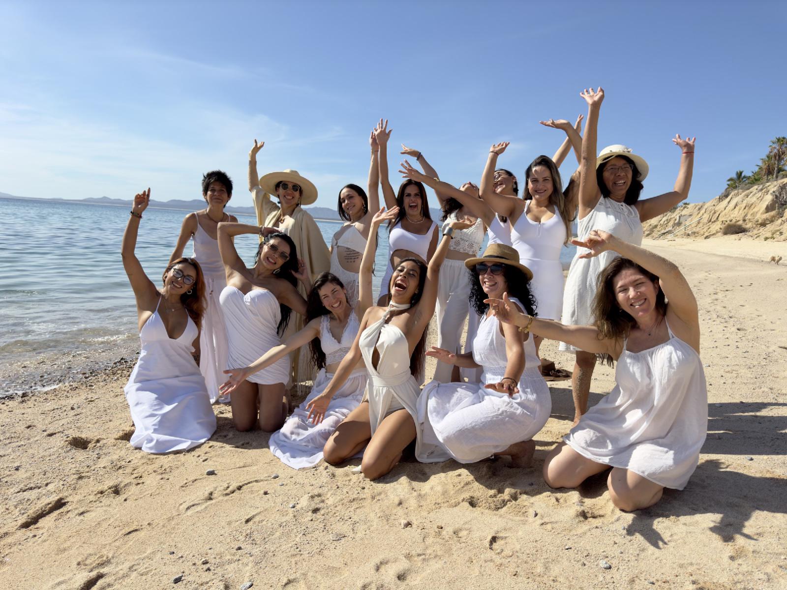 Un grupo de mujeres vestidas de blanco celebra en la playa con los brazos en alto, bajo un cielo azul despejado.