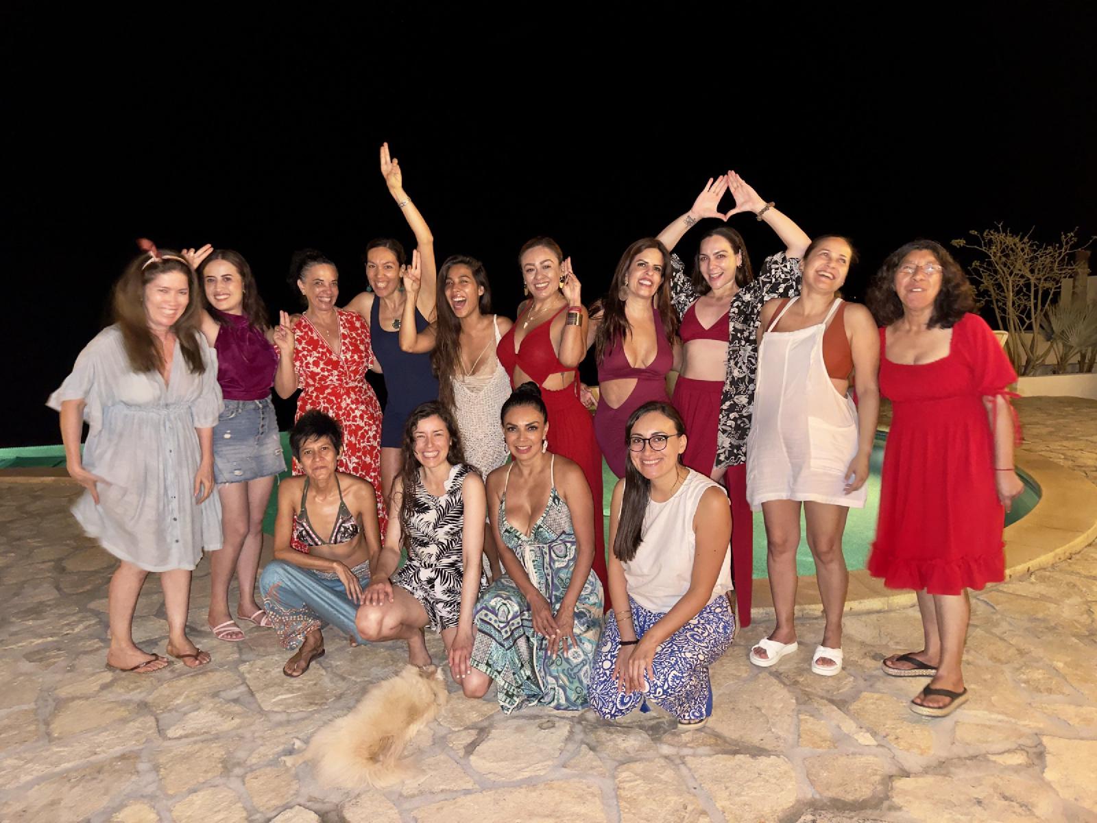 Foto grupal nocturna de mujeres sonrientes junto a una alberca, con vestidos de colores y un perrito peludo en primer plano.
