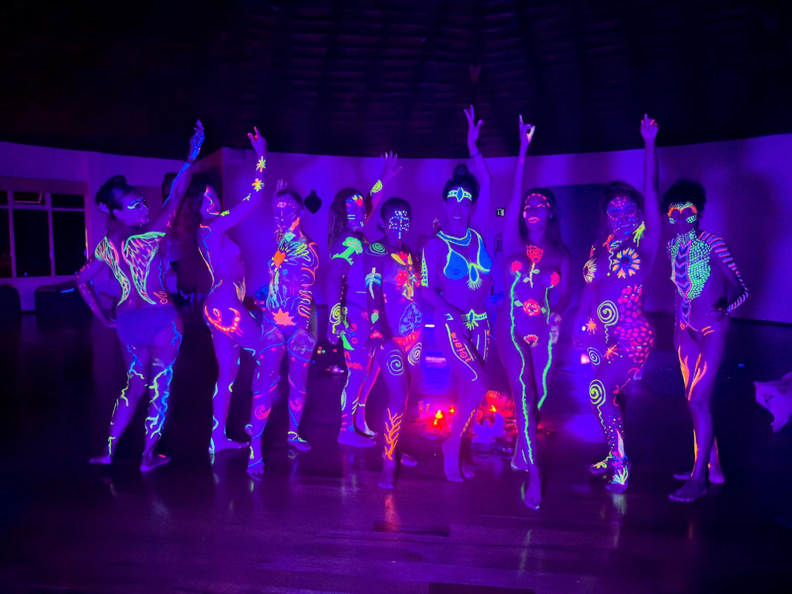 Mujeres con el cuerpo pintado con diseños fluorescentes posan bajo luz ultravioleta en una sala oscura, algunas con los brazos en alto.