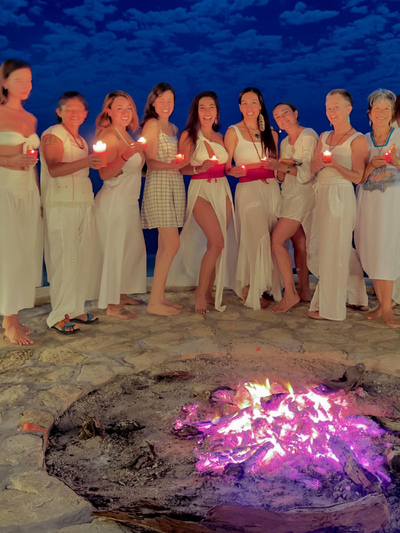 Mujeres vestidas de blanco sostienen velas rojas encendidas junto a una fogata en una terraza de piedra, frente al mar al anochecer.