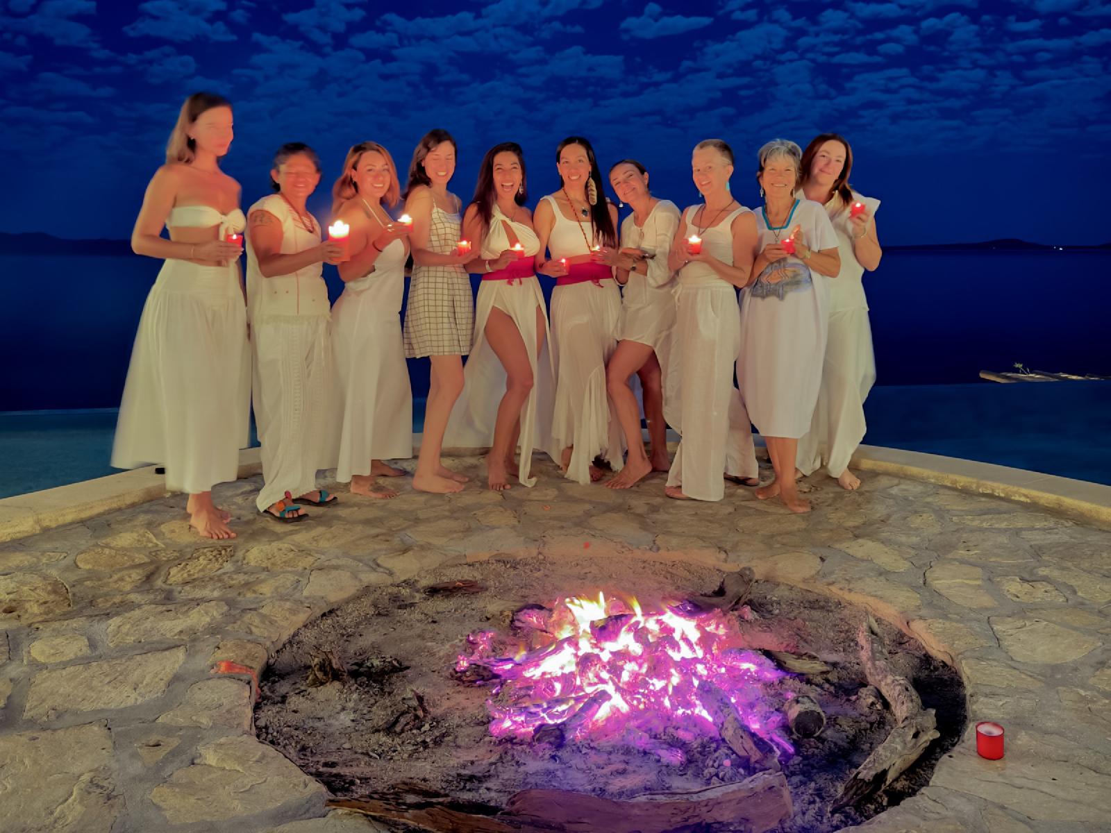 Mujeres vestidas de blanco sostienen velas rojas encendidas junto a una fogata en una terraza de piedra, frente al mar al anochecer.
