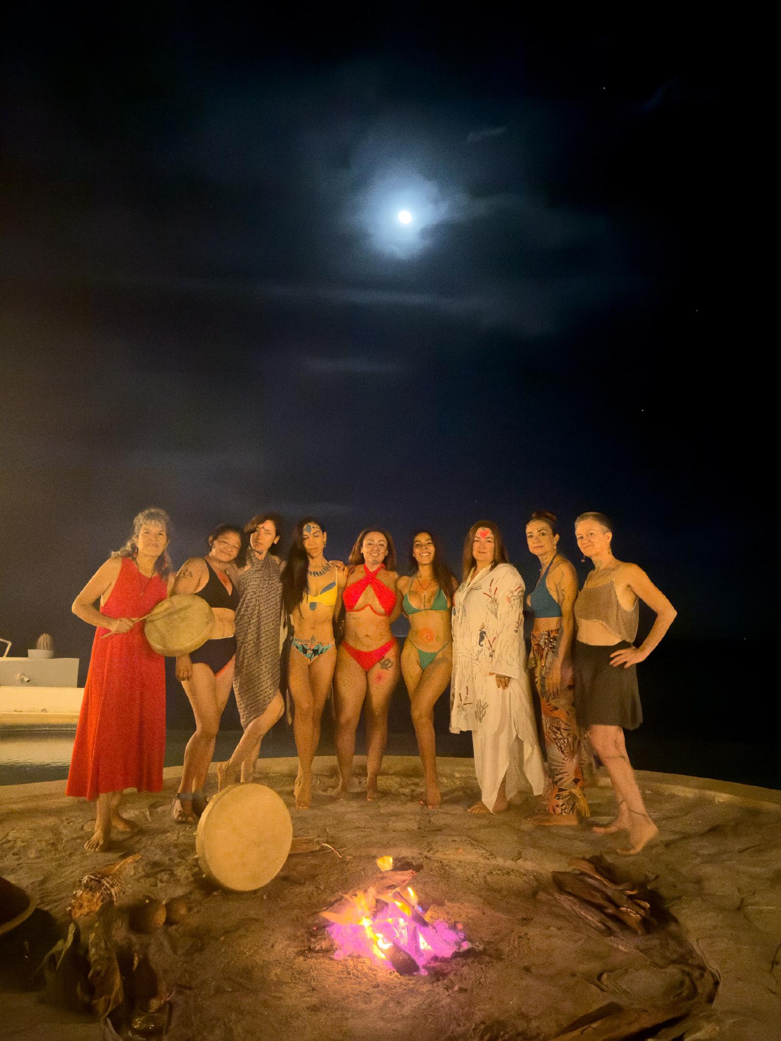 Mujeres, varias en traje de baño y con pintura en la piel, posan de noche alrededor de una fogata bajo la luna llena, con tambores.