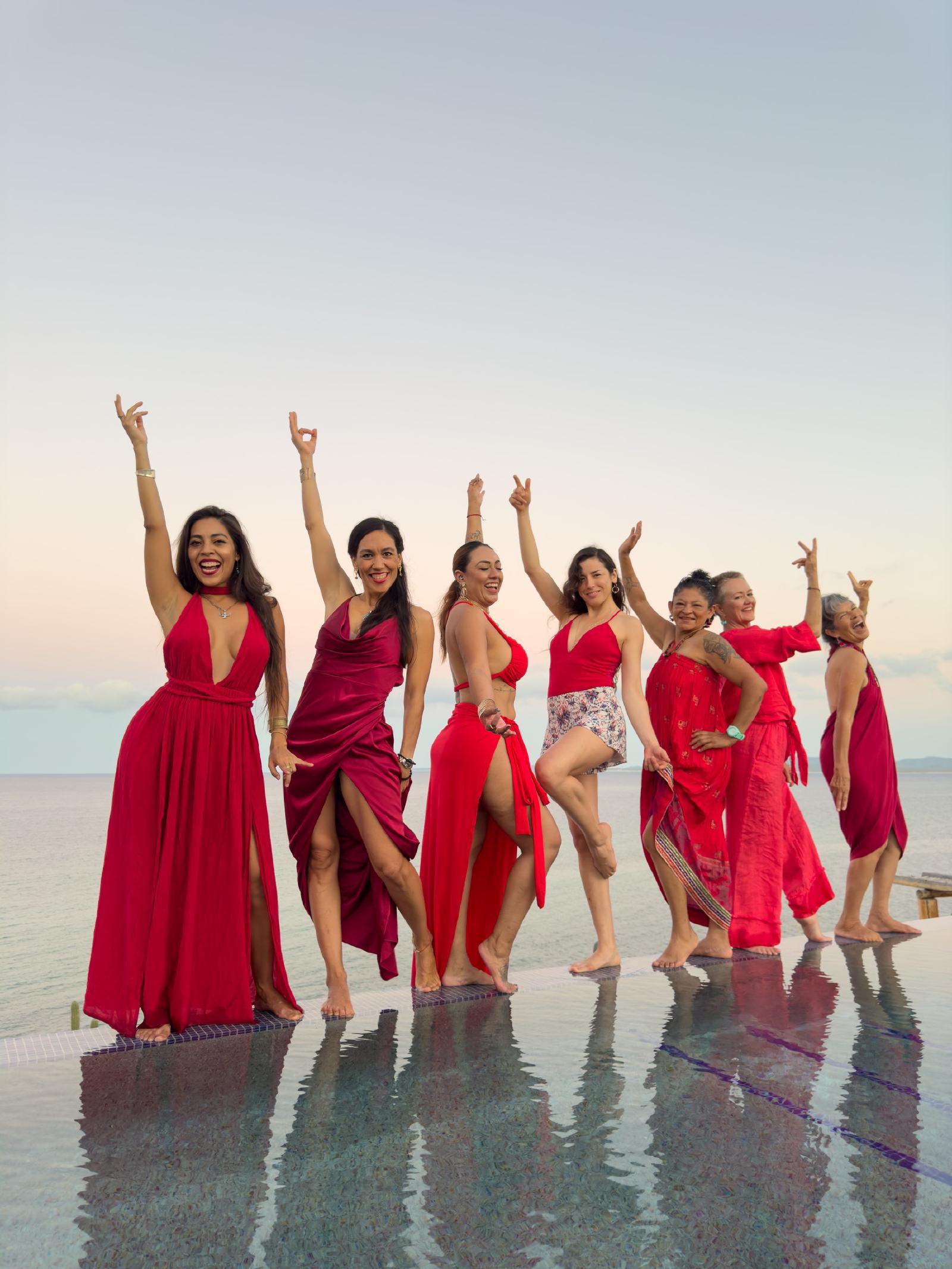 Siete mujeres vestidas de rojo bailan descalzas al borde de una alberca infinita frente al mar, al atardecer.