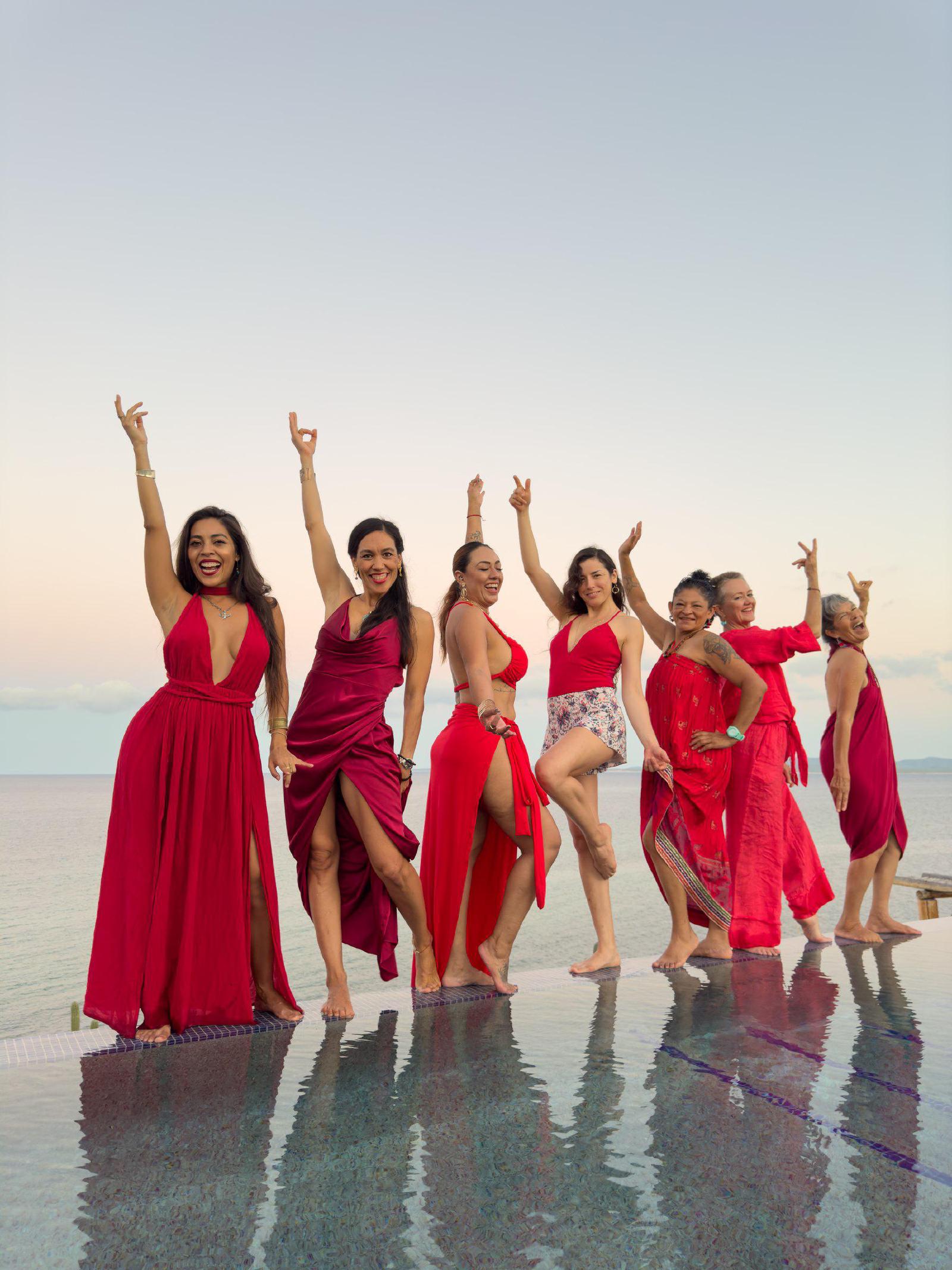 Siete mujeres vestidas de rojo bailan descalzas al borde de una alberca infinita frente al mar, al atardecer.